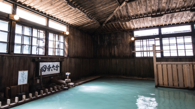 A serene indoor hot spring bath with wooden walls and ceiling, soft lighting, and milky blue water, giving the space a tranquil and rustic ambiance. This tattoo-friendly onsen welcomes all guests to experience ultimate relaxation.