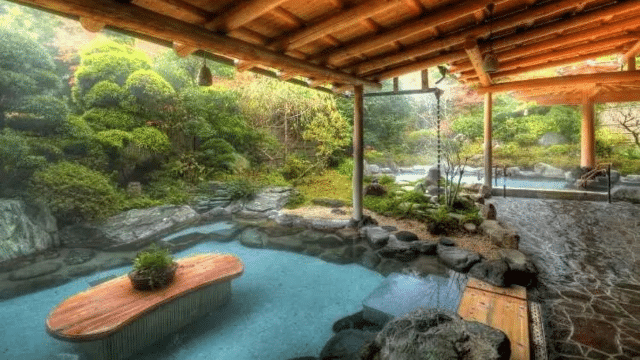 An outdoor, tattoo-friendly onsen with clear water, surrounded by lush greenery and covered by a wooden roof structure. A wooden platform sits in the water, while stone pathways wind around the pool.