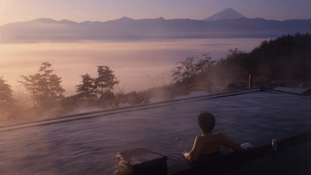 A person is soaking in a tattoo-friendly onsen during sunrise, with misty mountains and a distant peak in the background.