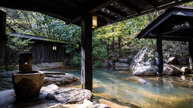 An outdoor tattoo friendly onsen sheltered by a wooden roof and surrounded by lush greenery, rocks, and a traditional Japanese building.
