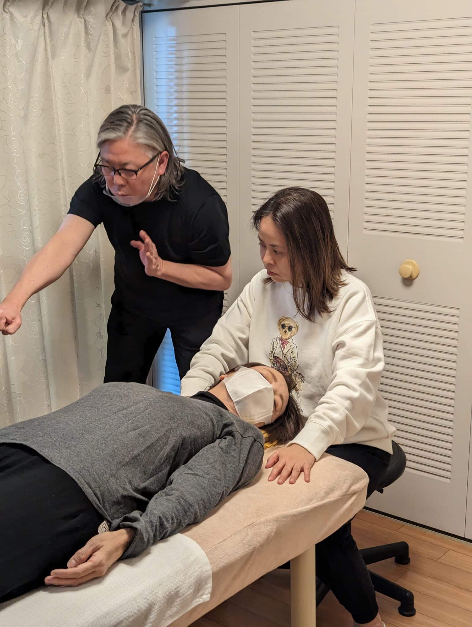 Two people perform a healing session: one lies on a table with a mask, receiving small face therapy, while the other two, seated and standing, place their hands above the body, focusing on providing therapeutic energy.