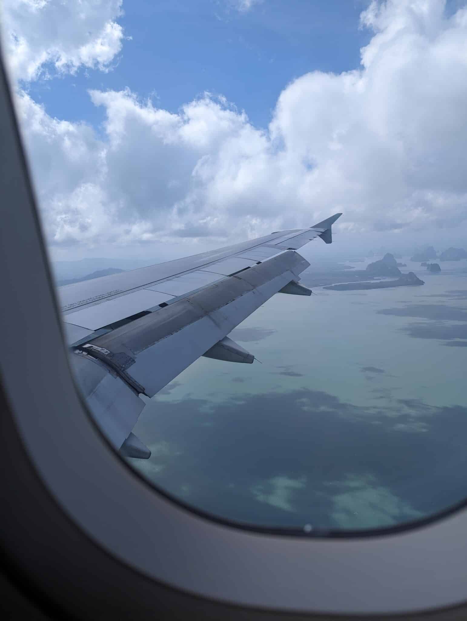 View of an airplane wing from a window seat, flying over a scenic landscape with blue sky, clouds, and a mix of land and water below