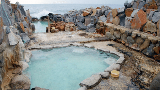 A natural hot spring with clear turquoise water is surrounded by rocky formations and overlooks the ocean in the distance, offering a welcoming atmosphere for everyone, including those seeking a tattoo-friendly onsen experience.