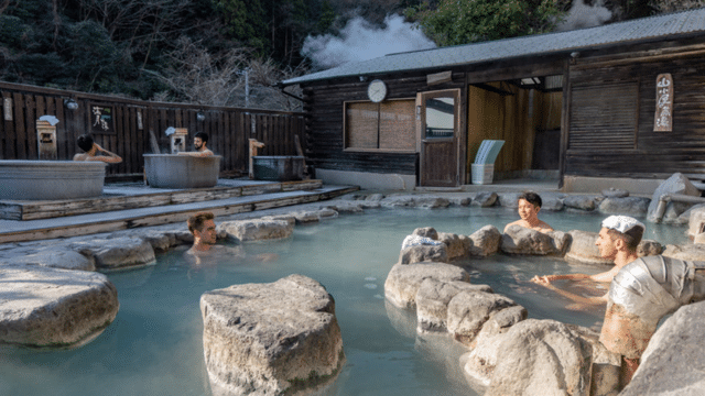 People relaxing in an outdoor hot spring with steam rising. Some are sitting in small tubs, while others are in a large central pool surrounded by rocks. A wooden building is in the background, highlighting this tattoo-friendly onsen's welcoming atmosphere for all visitors.