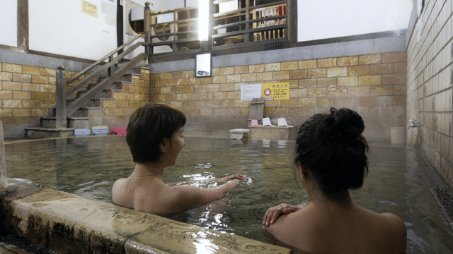 Two people relax in a steaming indoor hot spring bath, enjoying the warm embrace of the tattoo friendly onsen. The setting features tiled walls and wooden furniture, with signs visible on the wall in the background.