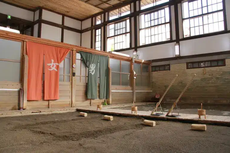 Traditional Japanese bathhouse entrance with red and green curtains labelled for women and men, next to a sand-filled sumo wrestling ring inside a wooden building—this tattoo friendly onsen welcomes all guests to experience authentic relaxation.