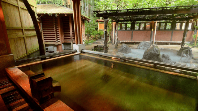 A serene Japanese onsen with wooden structures and clear hot spring water. The tattoo-friendly onsen is surrounded by green foliage and features both indoor and outdoor bathing sections, offering a welcoming retreat for all visitors.