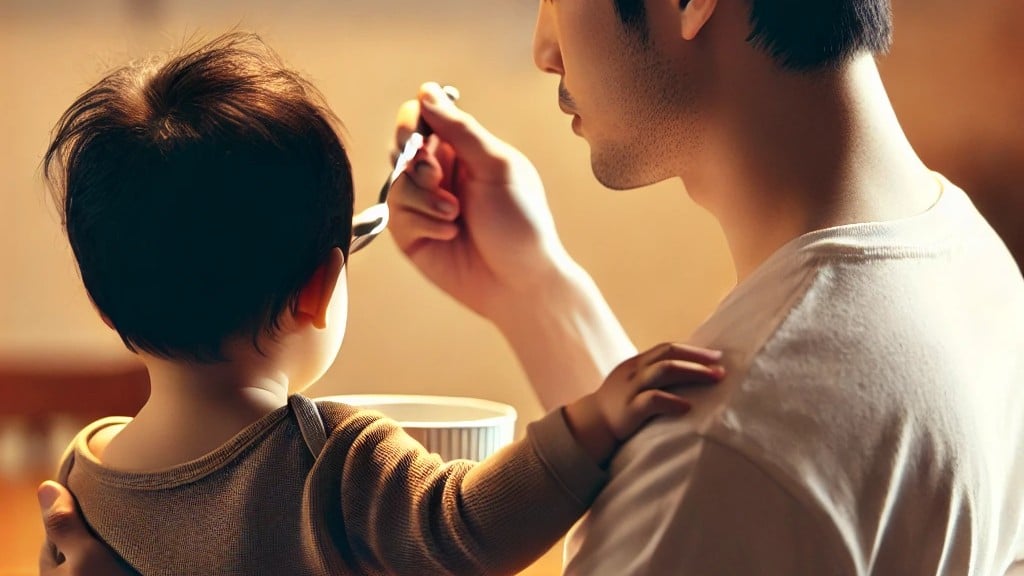 An adult is feeding a baby with a spoon, the little one nestled on their lap and holding onto them with one hand.