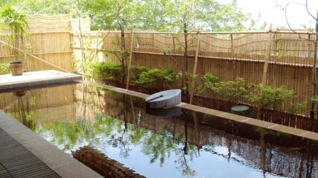 A tranquil, tattoo-friendly onsen with natural stone and foliage, surrounded by a bamboo fence, reflects trees and sky in its serene waters.