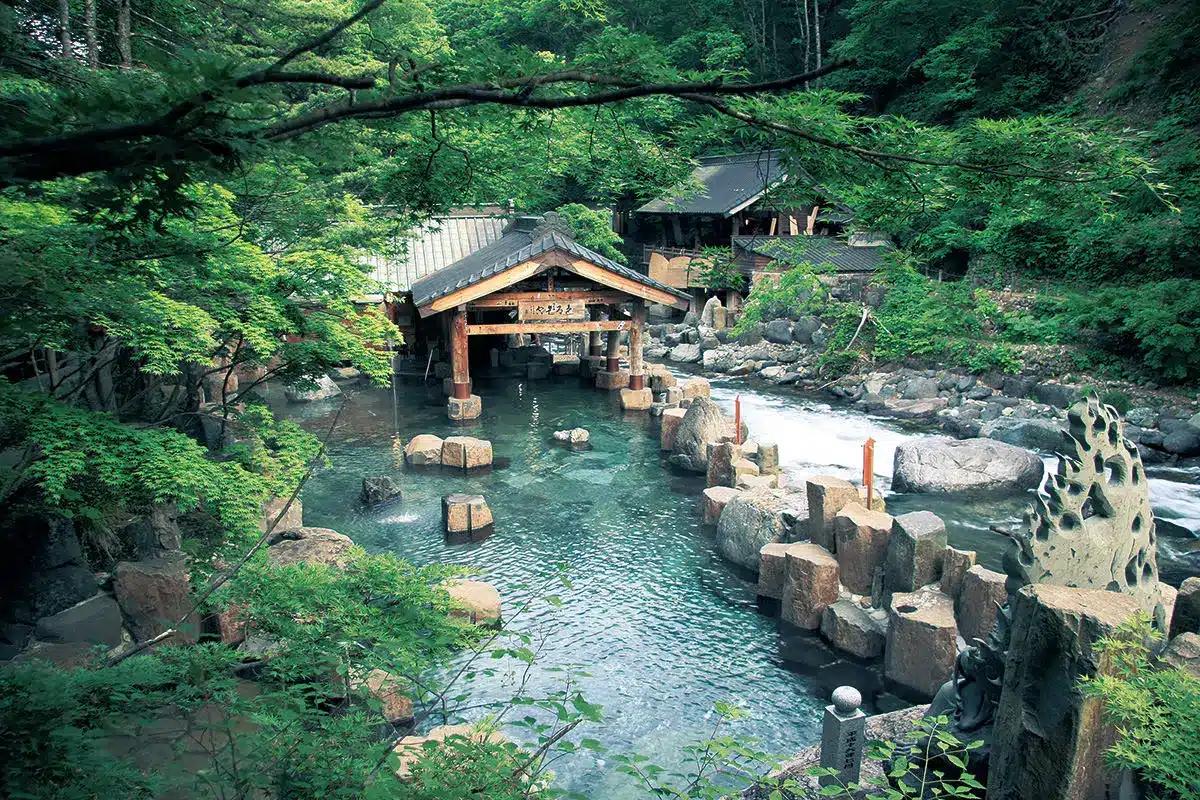 Outdoor Japanese onsen with a wooden pavilion, surrounded by green trees, rocks, and clear hot spring water in a tranquil natural setting—this tattoo friendly onsen welcomes all guests to unwind and enjoy the serene atmosphere.