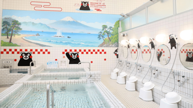A onsen features mural art depicting a coastal landscape and cartoon bear characters. Several washing stations with round mirrors and white stools are on the right side, accompanied by pools in the foreground.
