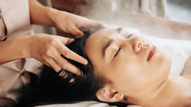 A person receives a head massage while lying down with eyes closed, a visible star tattoo behind their ear.