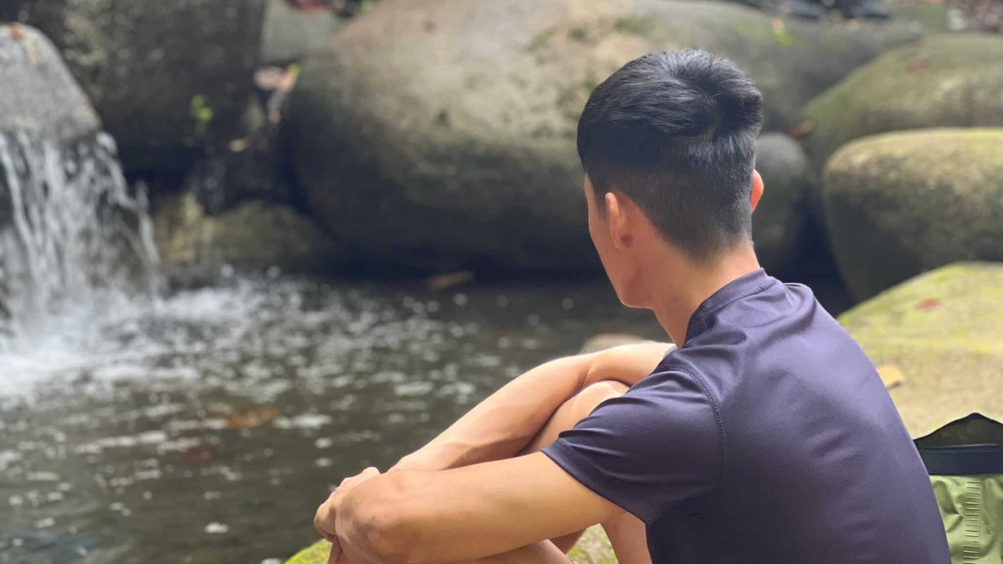 Two men are in nature near a small waterfall. One sits on a rock close to the water, facing away, while the other sits on a higher rock, facing the viewer. Lush greenery surrounds the scene, creating an ideal setting for forest bathing.