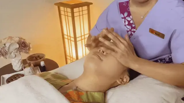A person reclines on a spa bed while a staff member in uniform pampers them with a soothing head massage and facial treatment. Spa products and elegant décor create a relaxing ambience in the background.