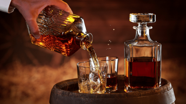 A hand pours amber liquid from a decanter into a glass on a barrel, with another full decanter and glass nearby.