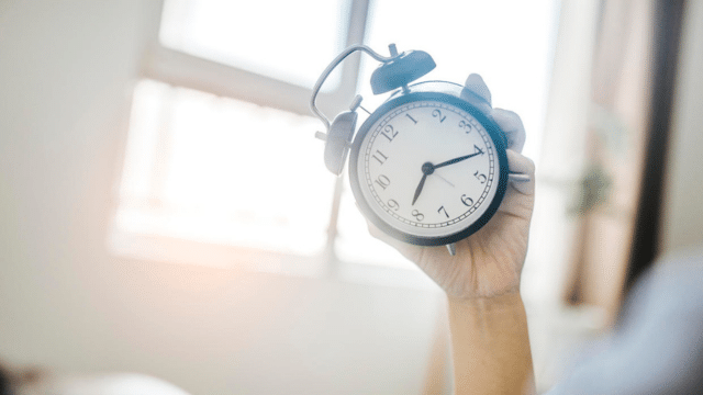 In a brightly lit room, a person holds a black analog alarm clock with a white face.