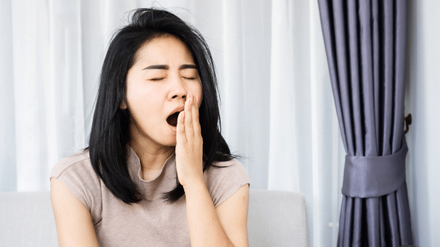 A person is sitting on a couch, yawning with their hand covering their mouth, perhaps pondering insomnia treatment. They have shoulder-length black hair and are wearing a light-colored shirt. A curtain is visible in the background.