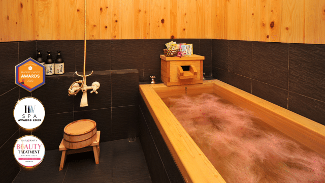 A wooden bathtub filled with water in a spa-like setting, surrounded by wooden and dark tiles. Nearby, awards and spa products are beautifully displayed, highlighting the sanctuary's dedication to tranquility and relaxation.