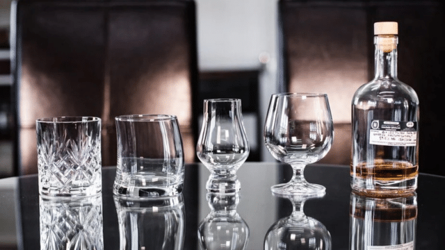 Four different types of empty glasses next to a partially filled liquor bottle on a reflective black surface.