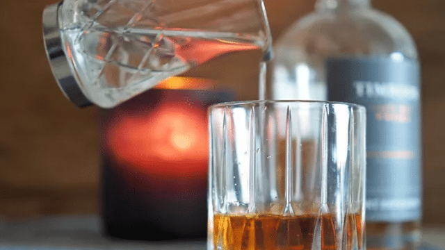 A glass is being filled with whiskey from a jigger, with a bottle and a lit candle in the background.