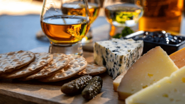 A cheese platter with assorted cheeses, crackers, pickles, and a small glass of amber-colored liquid, likely whiskey.