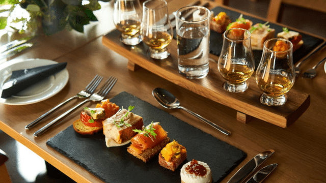 A gourmet meal with appetizers on slate plates and glasses of whiskey on a wooden tray, set on a wooden table.