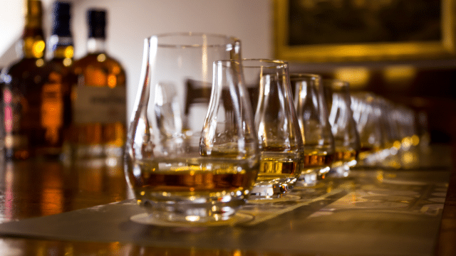 A row of whiskey glasses with varying amounts of amber liquid lined up on a table, with several whiskey bottles blurred in the background.