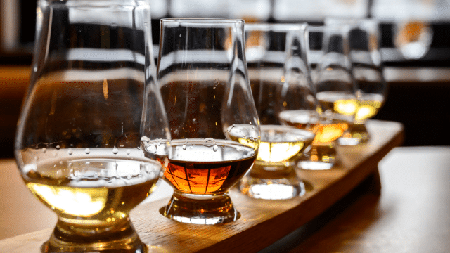 A row of whiskey glasses filled with different shades of whiskey on a wooden tray, set on a table.