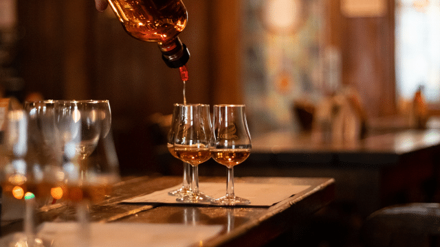 A person pours whiskey into two glasses on a wooden table in a dimly lit bar.