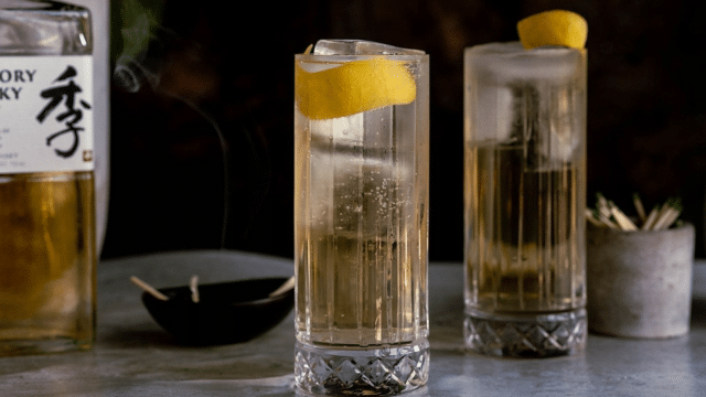 Two highball glasses filled with a clear bubbly drink, topped with lemon twists, sit on a table. A bottle of Japanese whisky is nearby along with a bowl with incense and toothpicks.