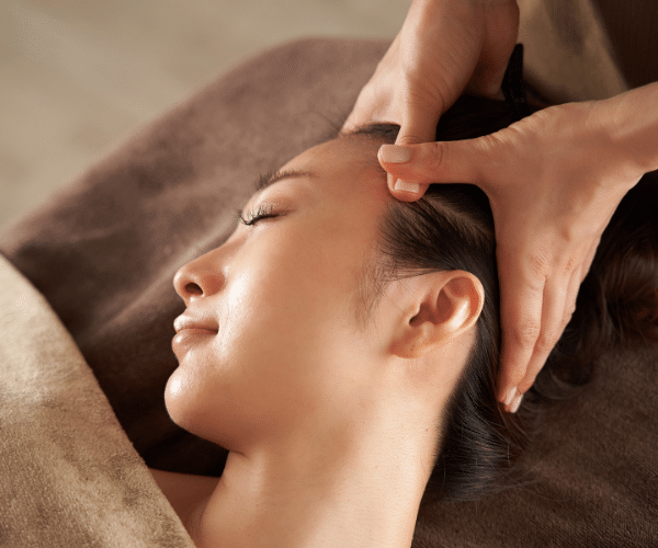 Lying comfortably on a towel-covered surface, eyes gently closed, the person relishes the soothing sensations of a head massage.