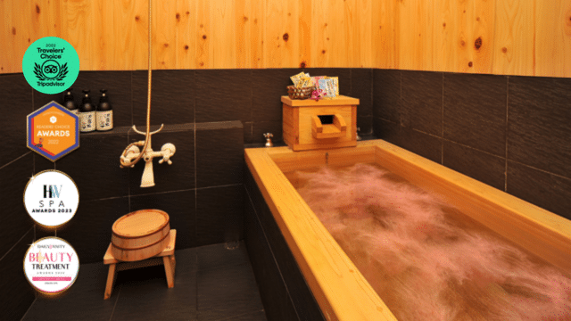 A luxurious wooden bathtub filled with water, surrounded by dark tiles and bath products. Various award badges are displayed in the top left corner.
