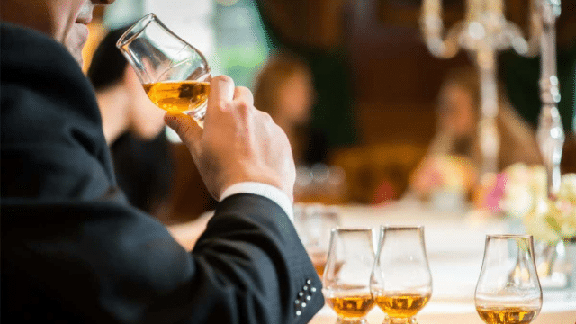 A person in a suit sniffs a glass of amber liquid, with three additional filled glasses on a table in a well-lit room.