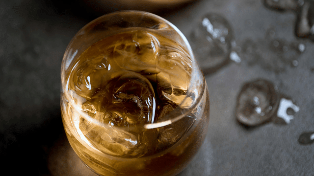 A glass filled with a brown liquid and ice cubes on a dark surface; scattered ice cubes are visible in the background.