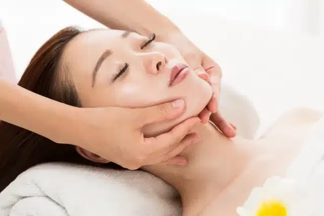 A person lying on a towel receives a facial massage, with hands gently holding their chin and jawline for small face correction.
