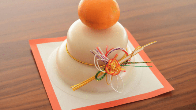 Traditional Japanese kagami mochi with two stacked rice cakes, a small orange on top, and colorful decorative threads evoke a festive spirit reminiscent of Japanese celebrations, elegantly placed on a wooden surface.