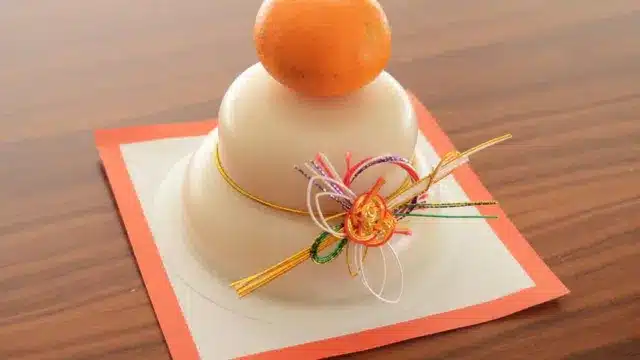 A traditional Japanese kagami mochi with two stacked rice cakes, an orange on top, and decorative cord, placed on a square paper mat—often displayed in Japan and sometimes during Chinese New Year celebrations.