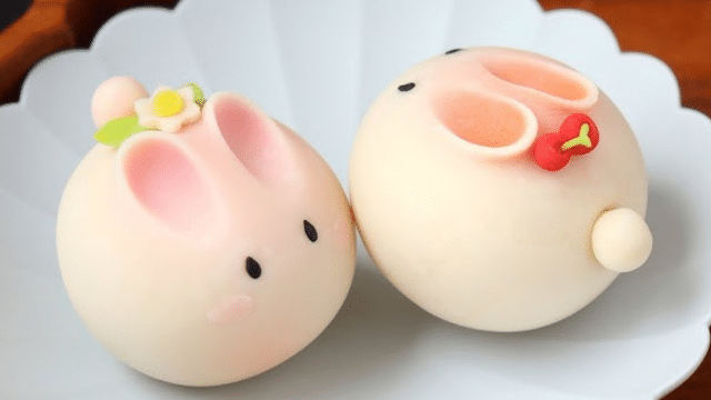Two round, bunny-shaped steamed buns with pink ears sit on a white plate. One has a small flower, while the other sports a red bow on the back, beautifully blending traditions from Japan with a festive touch.