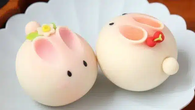 Two round Japanese sweets shaped like rabbits with pink ears and small decorative flowers, placed on a white plate, evoke a festive charm reminiscent of Chinese New Year celebrations in Japan.