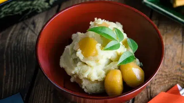 A red bowl containing mashed sweet potatoes topped with cooked yellow chestnuts and garnished with green leaves, placed on a wooden surface—a festive dish enjoyed during Chinese New Year in Japan.