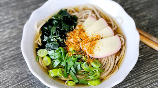 A bowl of soba noodles with sliced kamaboko, seaweed, green onions, and sprinkled seasoning sits on a wooden table with chopsticks.