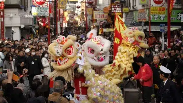 A crowded street scene in Japan shows a traditional lion dance performance with two lion costumes and many onlookers during Chinese New Year celebrations.