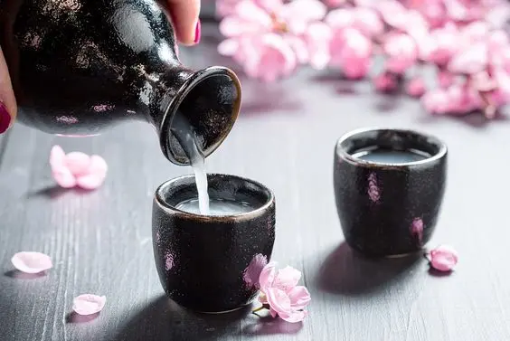 A hand pours cloudy sake from a black ceramic bottle into a matching cup, with pink cherry blossoms scattered on a dark table—a scene reminiscent of the cherry blossom forecast season.