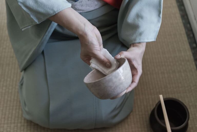 A person in a blue kimono gracefully conducts a Japanese tea ceremony, carefully preparing a tea bowl on a tatami mat, with the traditional utensil poised nearby.