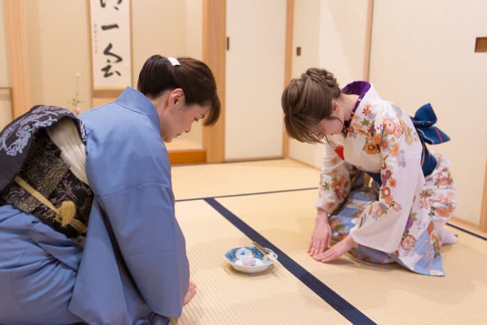 Two people in traditional Japanese attire are performing a Japanese tea ceremony, kneeling gracefully on tatami mats with a tea bowl between them.