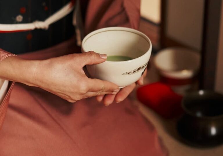 A person wearing a pink kimono is elegantly holding a white bowl with matcha, embodying the serene ritual of a Japanese tea ceremony in a traditional setting.