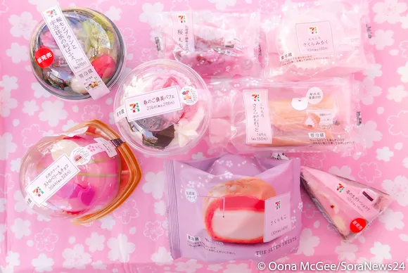 Assorted cherry blossom-themed Japanese desserts in clear and pastel pink packaging, arranged on a pink floral background, perfect for celebrating the season during the cherry blossom forecast.