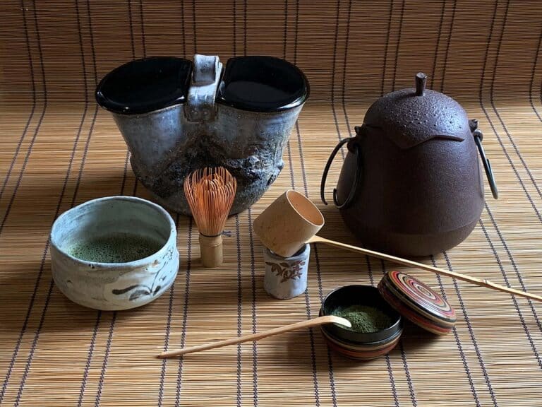 Experience the elegance of a Japanese tea ceremony with this exquisite set featuring a teapot, two bowls, bamboo whisk, ladle, tea caddy, and bamboo scoops, all beautifully arranged on a bamboo mat background.