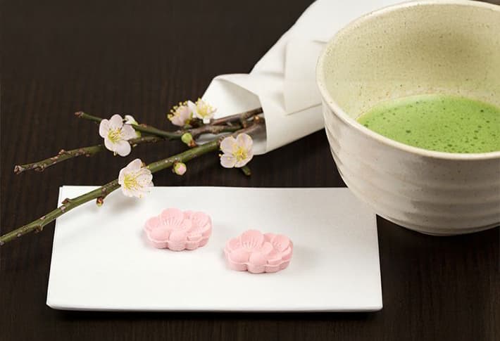 A bowl of green tea, reminiscent of a Japanese tea ceremony, sits beside a white plate adorned with two pink flower-shaped sweets. Delicate cherry blossoms grace the napkin in the background.