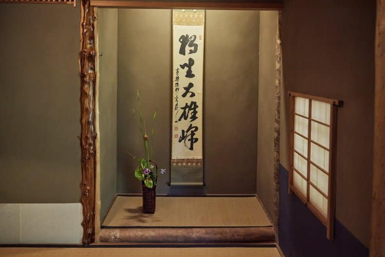 A serene Japanese tokonoma features a scroll displaying calligraphy and a delicate floral arrangement in a vase on the tatami mat floor, evoking the tranquility of a traditional Japanese tea ceremony.